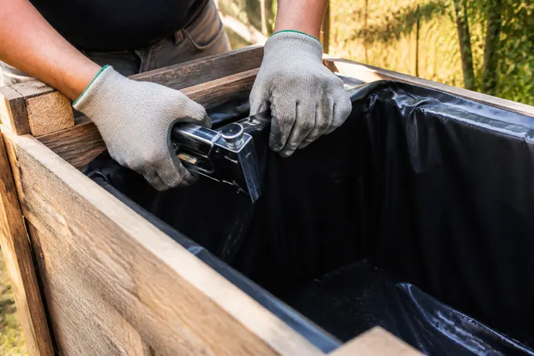Des mains gantées fixent une bâche plastique à l'intérieur d'une jardinière en palettes, assurant ainsi une première couche d'étanchéité essentielle.