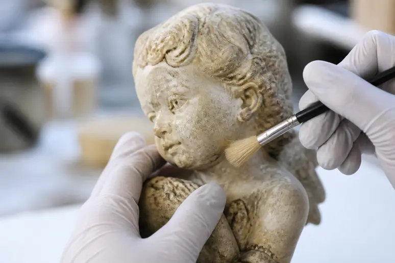Mains gantées d'un restaurateur nettoyant délicatement une sculpture en albâtre, montrant le savoir-faire de la pierre.